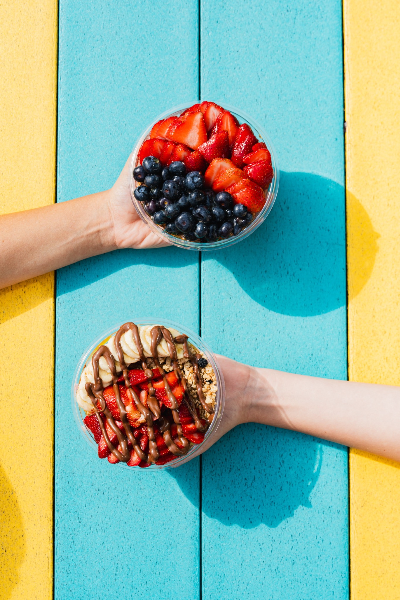Two Playa bowls held over a turquoise and yellow table, topped with berries and banana